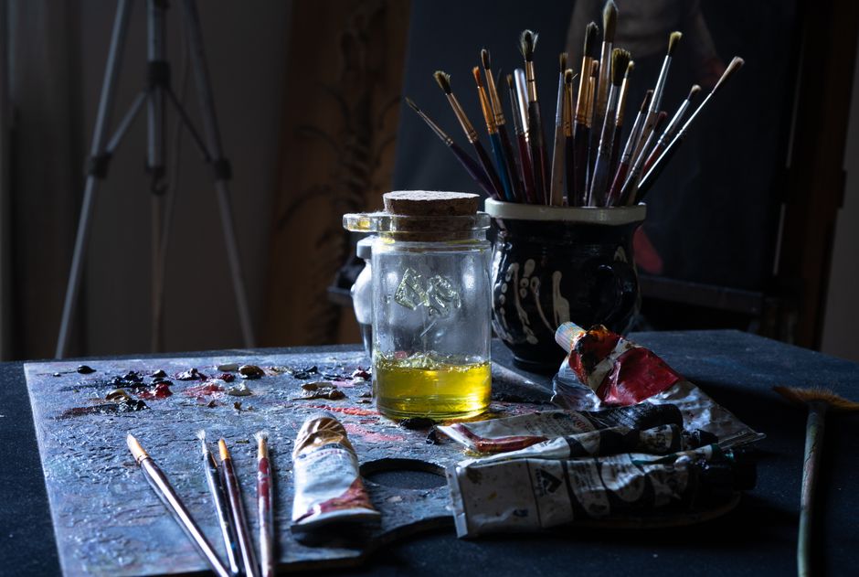 A cluttered artist's table filled with paint tubes, brushes, and a glass jar.