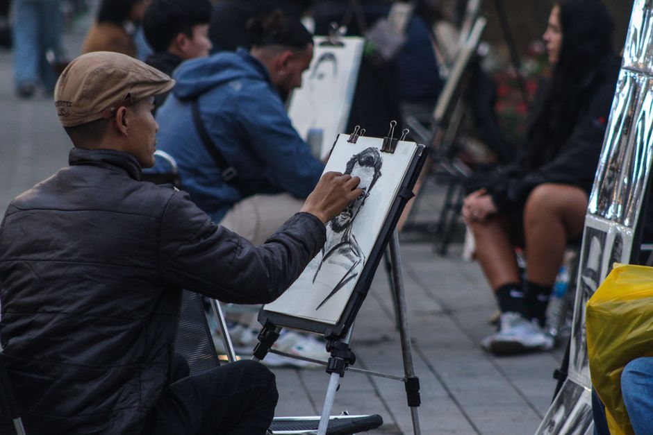 A street artist creates a detailed charcoal portrait sketch outdoors with surrounding onlookers.